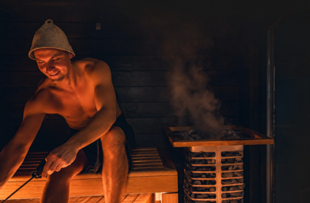 A man in a wooden sauna wearing a wool sauna hat, steam rising from the stove